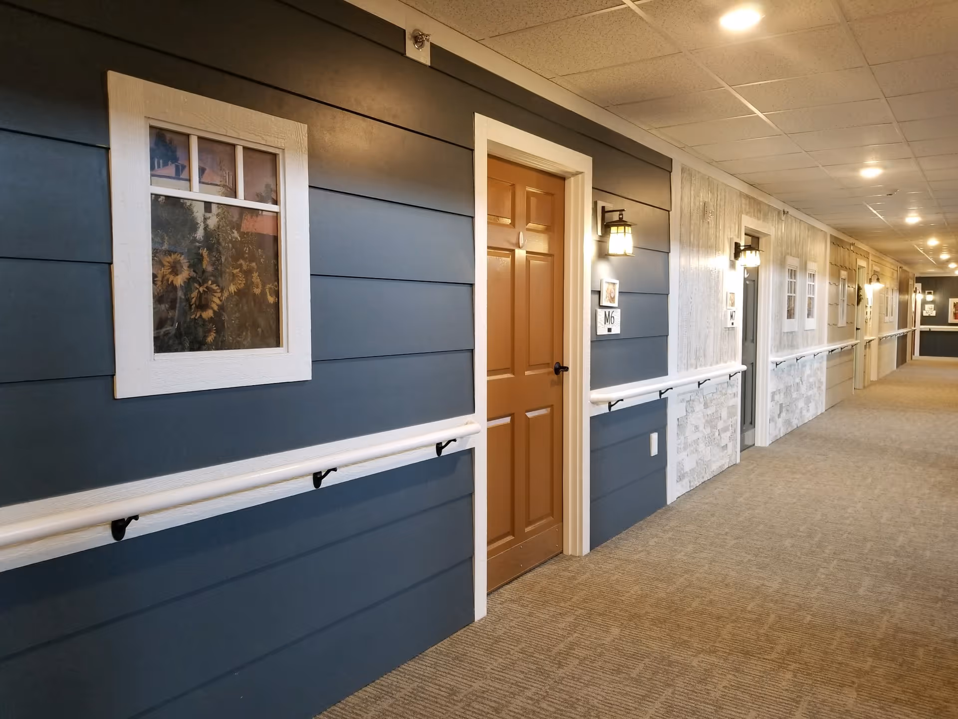 Carpeted interior hallway of a senior living facility with doors, handrails, wall lanterns and decorative windows.
