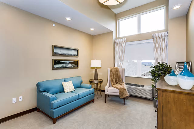 Small furnished sitting room with a teal loveseat, upholstered armchair with throw, side table and lamp, dresser with decorative vases, and a large window.
