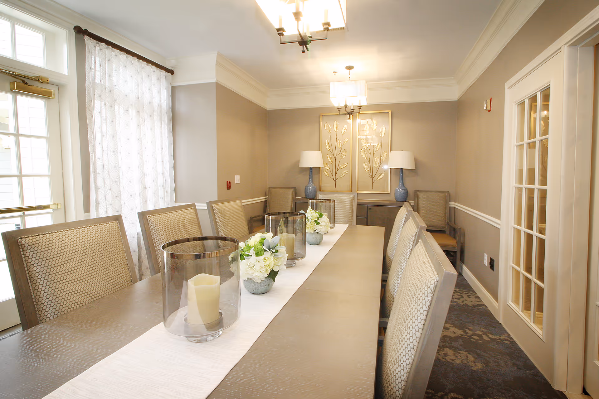 A well-lit dining room with a long rectangular table surrounded by cushioned chairs. The table is decorated with a white runner, glass candle holders with candles, and small flower arrangements. The room has beige walls, two blue lamps on a sideboard, and framed gold leaf artwork on the wall. There is a window with sheer white curtains and a glass door with multiple panes on the right side.
