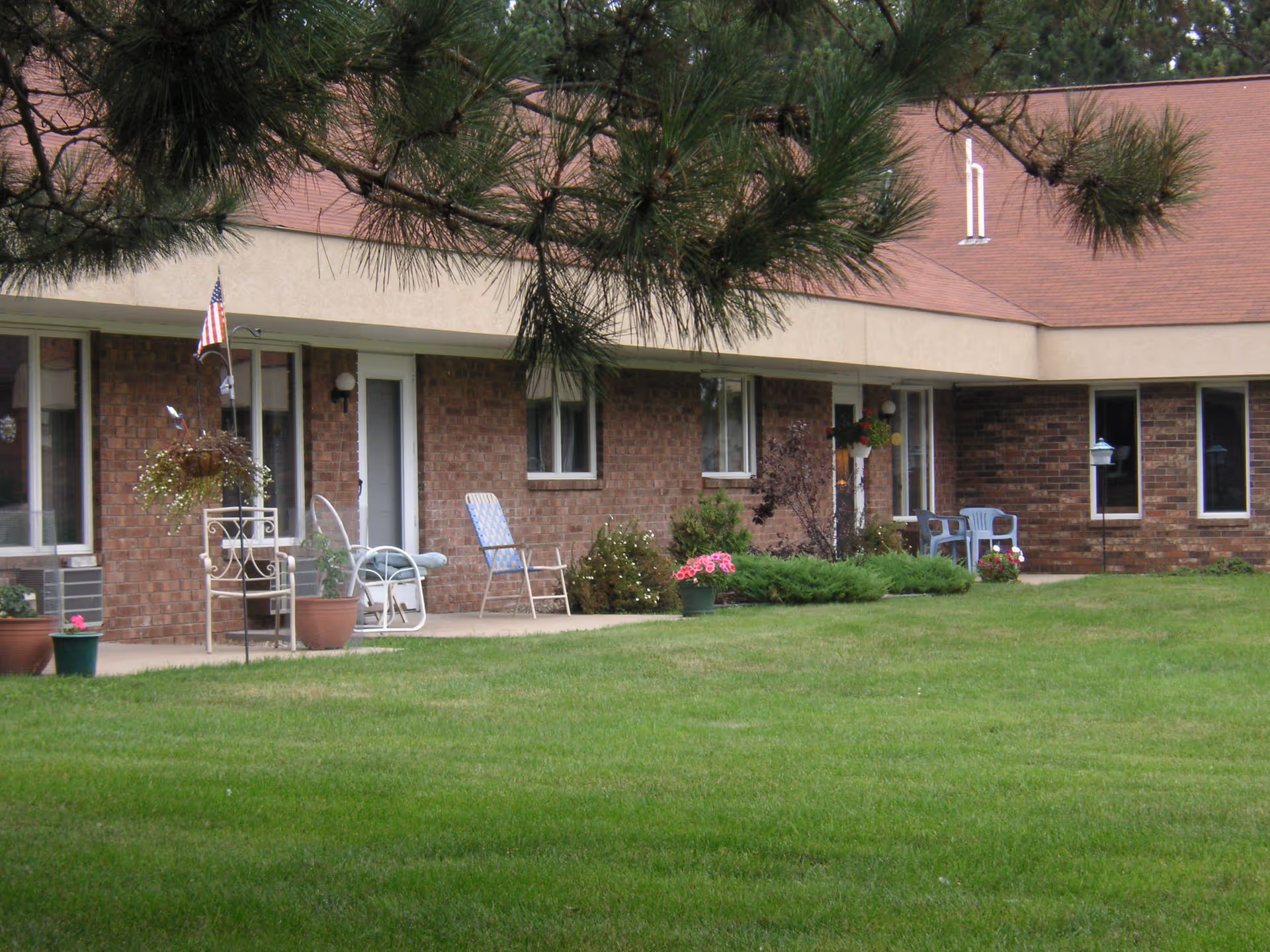 Exterior view of a single-story brick building with a sloped roof, featuring several doors and windows. There are chairs, potted plants, and an American flag on the patio area in front of the building. Green grass and some bushes are visible in the foreground, with tree branches partially framing the top of the image.