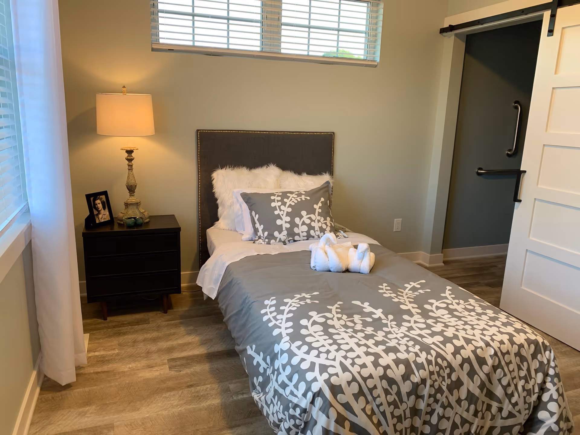 Small bedroom with a single bed dressed in gray patterned bedding, a nightstand with a lamp and framed photo, and a sliding barn door.