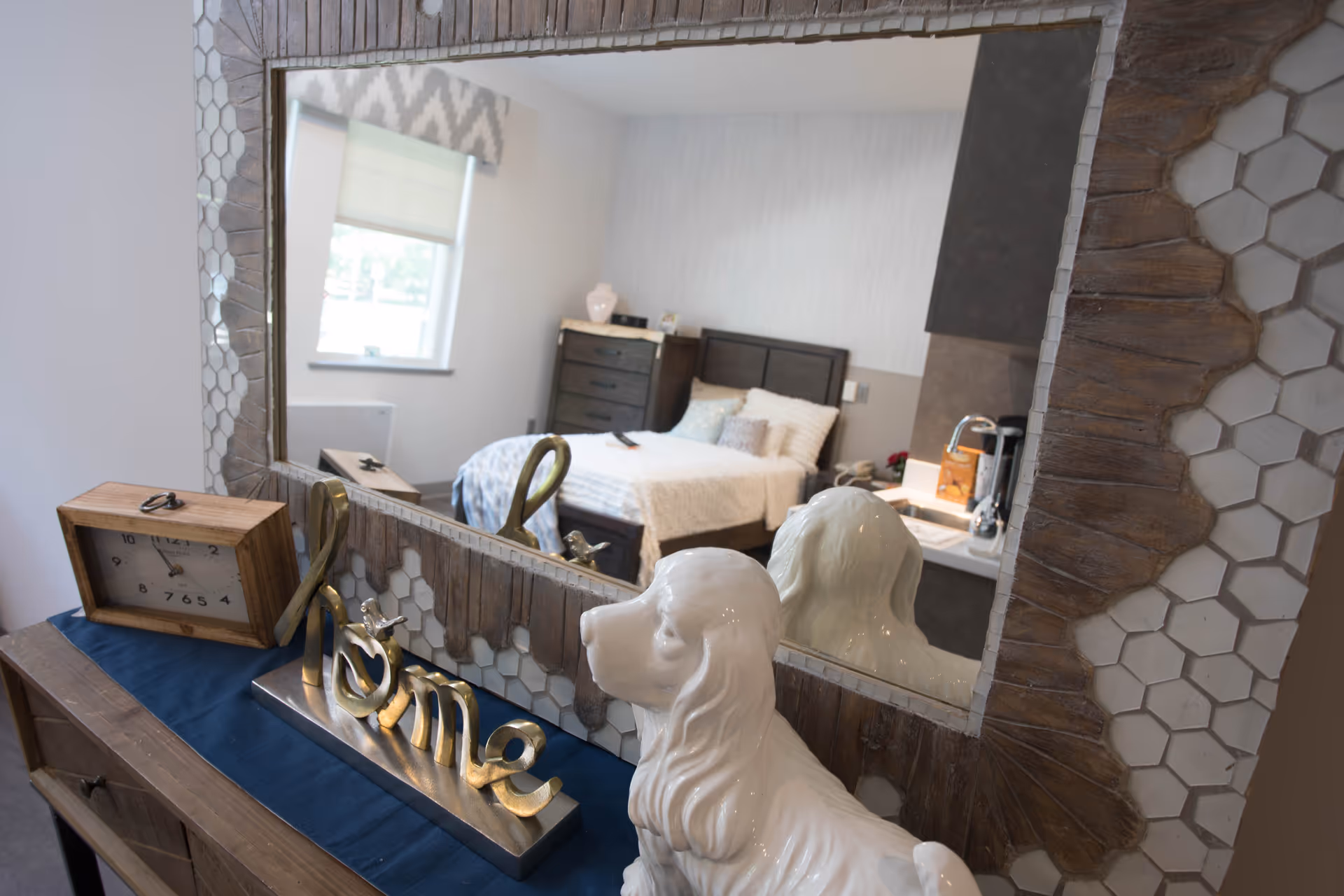 A decorative mirror with a wooden and hexagonal tile frame reflects a bedroom with a bed, dresser, and window. In front of the mirror is a wooden clock, a decorative sculpture spelling 'Home', and a white ceramic dog figurine on a wooden table with a blue runner.