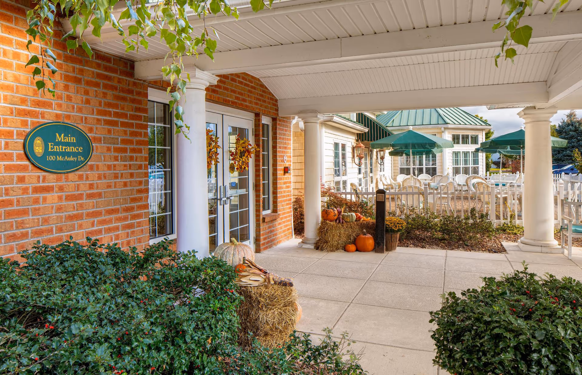 Covered entrance area of a brick building with white columns and glass double doors decorated with autumn wreaths. There are hay bales, pumpkins, and fall decorations near the entrance. In the background, there is an outdoor patio area with white chairs and tables under green umbrellas, surrounded by a white picket fence.