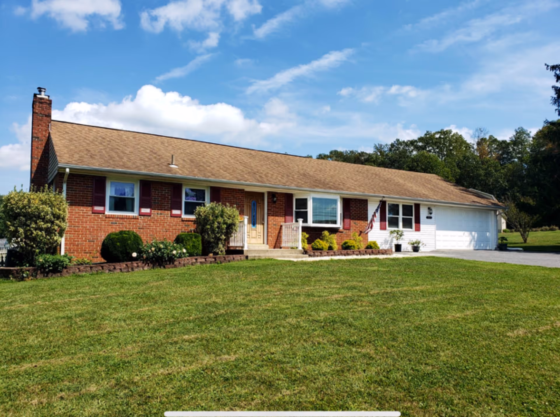 Single-story brick house with a brown shingled roof, white garage door, and a front porch with steps. The house is surrounded by a well-maintained lawn and some bushes. The sky is blue with some clouds.