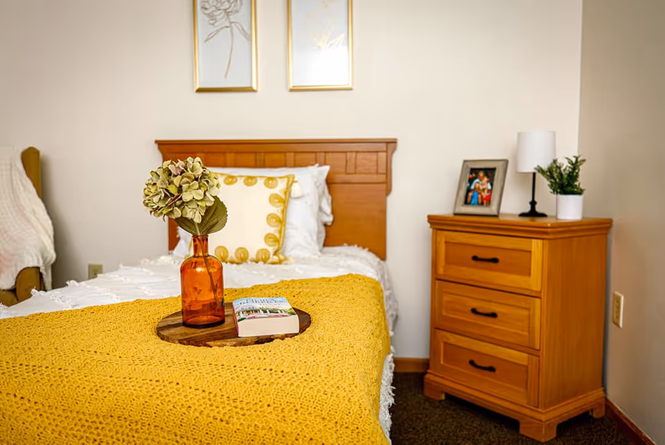 A cozy bedroom with a wooden headboard bed covered in white bedding and a yellow knitted blanket. On the bed is a wooden tray holding an amber glass vase with flowers and a book. Next to the bed is a wooden nightstand with three drawers, a small lamp, a framed photo, and a potted plant. Two framed floral artworks hang on the wall above the bed.