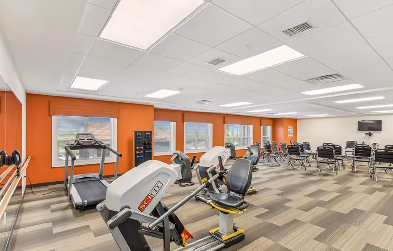 A spacious fitness room with exercise equipment including treadmills, stationary bikes, and weight racks. The room has large windows with orange walls and a light-colored patterned floor. There are several chairs arranged in rows facing a wall-mounted TV.