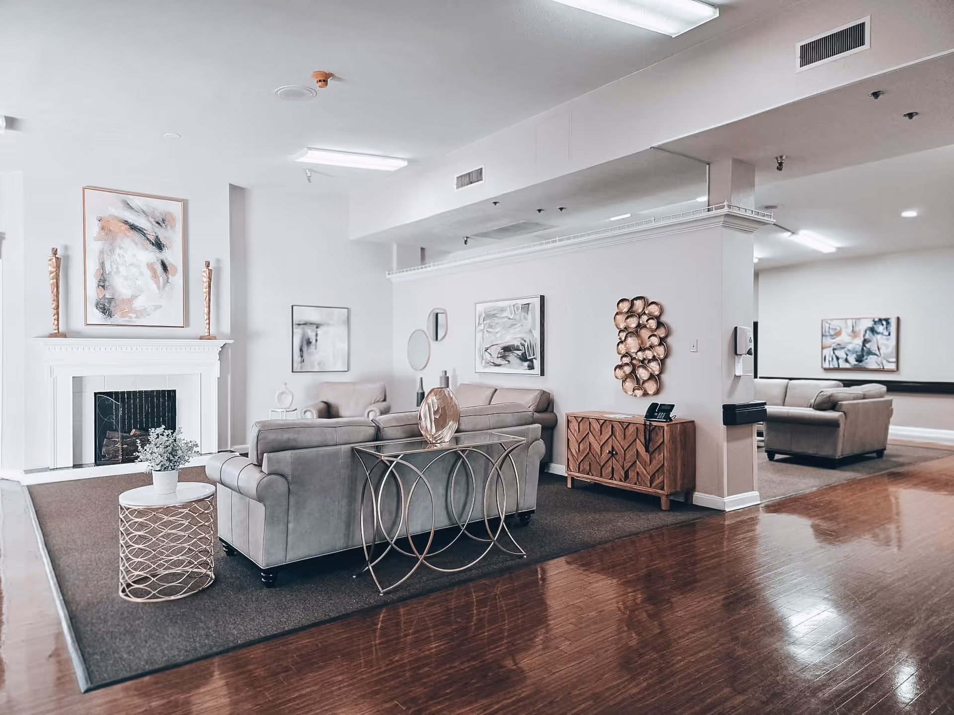 A spacious living room area with light gray sofas arranged around a white fireplace. The room features modern decor including abstract paintings, a decorative wooden sideboard, a glass-top console table, and a small round side table with a plant. The floor is polished wood with a large area rug under the seating area. The walls are painted white, and the ceiling has recessed lighting.