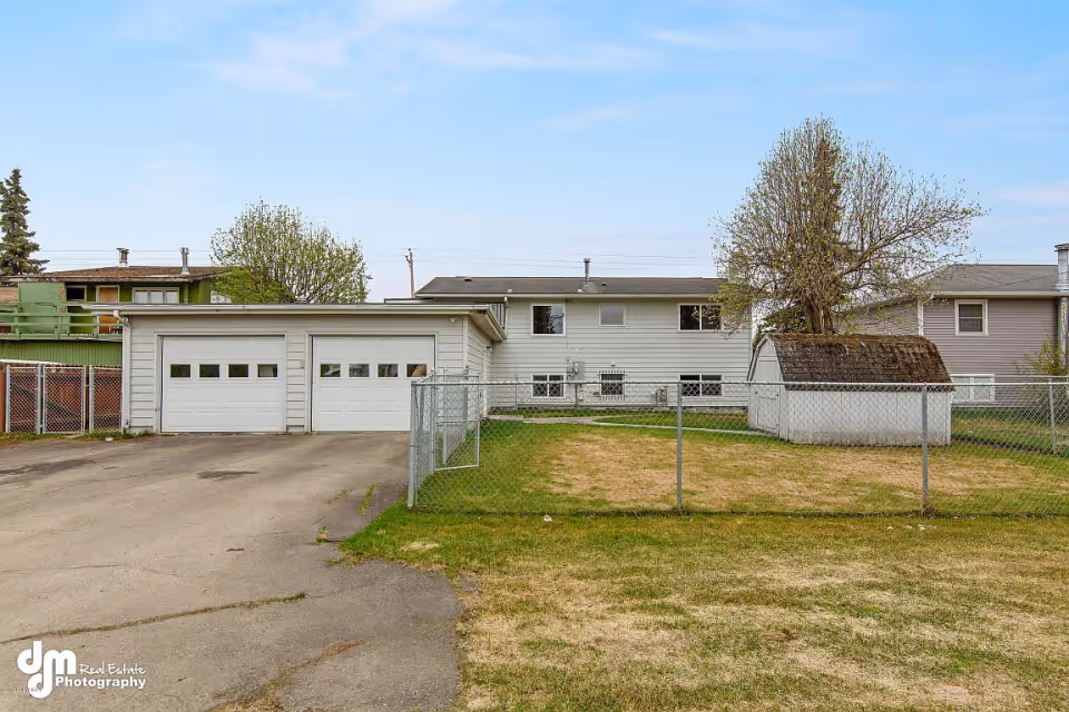 Exterior view of a residential building with a two-car garage, a fenced yard, and a small shed. The grass is patchy and there are a few trees around the property under a partly cloudy sky.