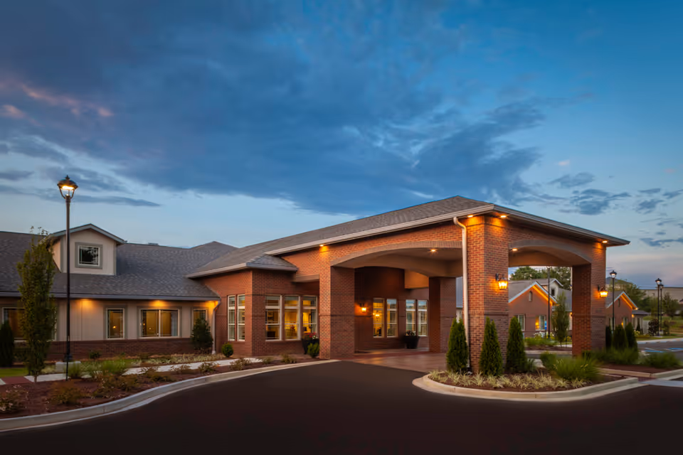 Exterior view of a single-story assisted living facility building with brick and beige siding, featuring a covered entrance with warm lighting and landscaped surroundings under a partly cloudy evening sky.