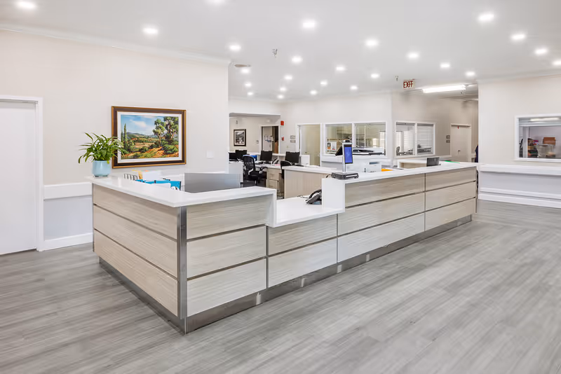 A modern reception desk area in a senior living facility with light wood paneling and white countertops. The space is well-lit with recessed ceiling lights and features a framed landscape painting on the wall, a potted plant, and office equipment behind the desk. The flooring is a light gray wood-style material.