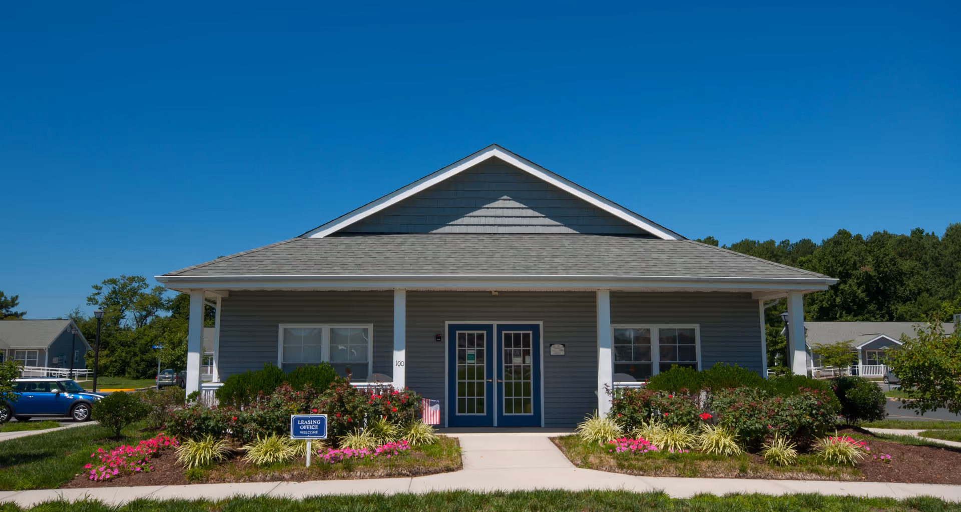 Exterior view of a single-story building with gray siding and a gabled roof under a clear blue sky. The building has a covered porch with white pillars, double blue doors with glass panes, and windows on either side. There are well-maintained flower beds with pink flowers and green shrubs in front of the building, along with a small sign that reads 'Leasing Office Welcome'. A blue car is parked on the left side, and other similar buildings and trees are visible in the background.