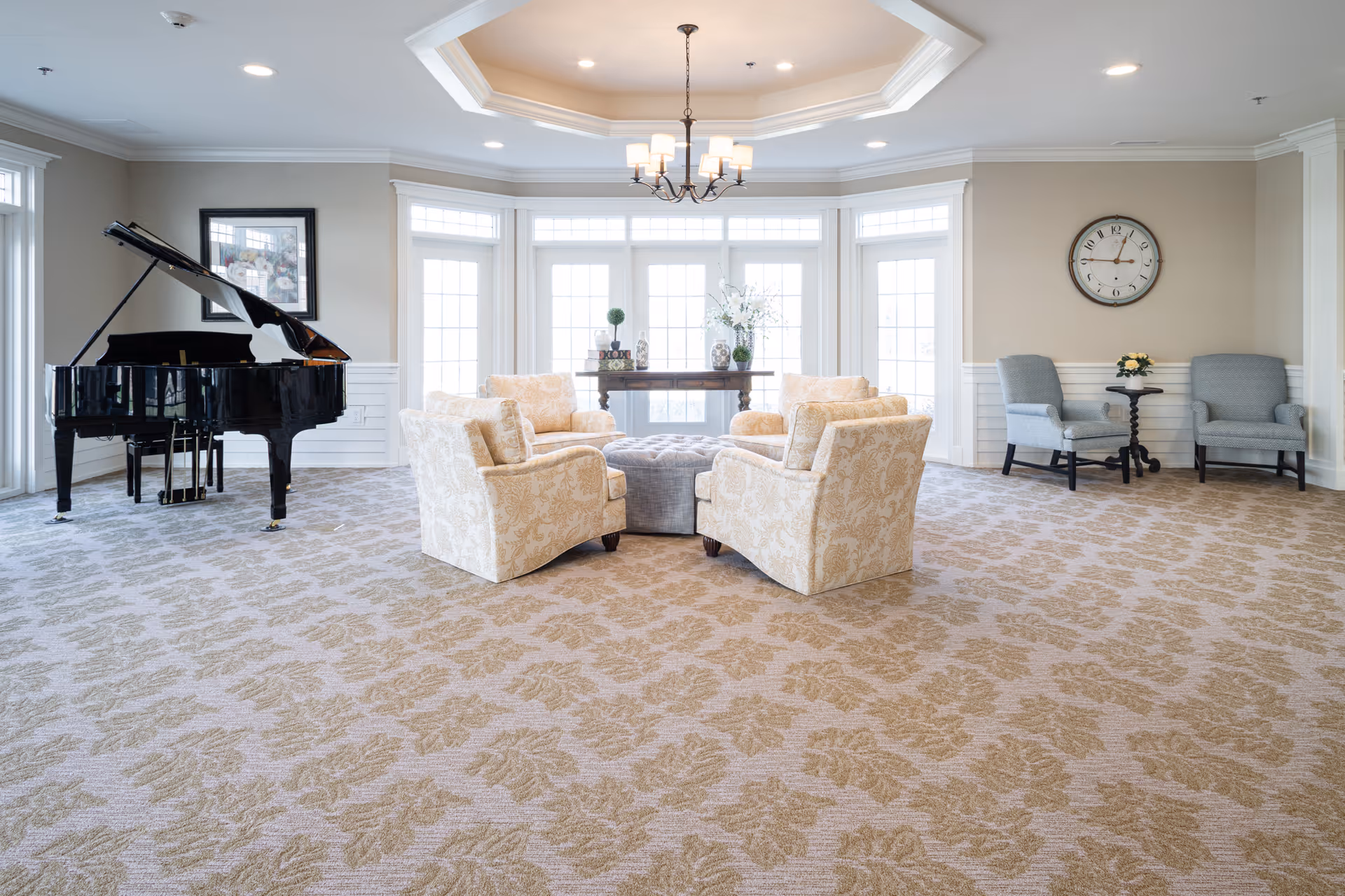 A spacious, well-lit living room with a patterned beige carpet, four upholstered armchairs arranged around a central ottoman, a grand piano to the left, a chandelier hanging from a tray ceiling, large windows with white frames letting in natural light, a small table with decorative items, two additional armchairs with a small round table between them, and a wall clock.