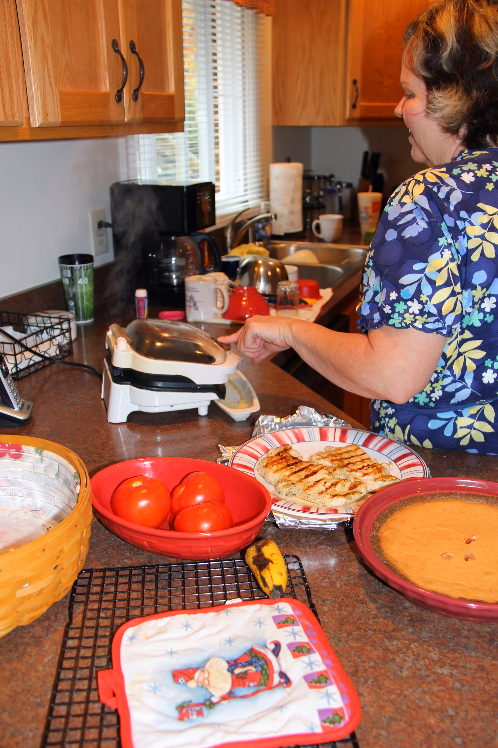 A person at a kitchen counter preparing food with a sandwich/waffle maker, plates of grilled chicken, a bowl of tomatoes, and a pie visible.