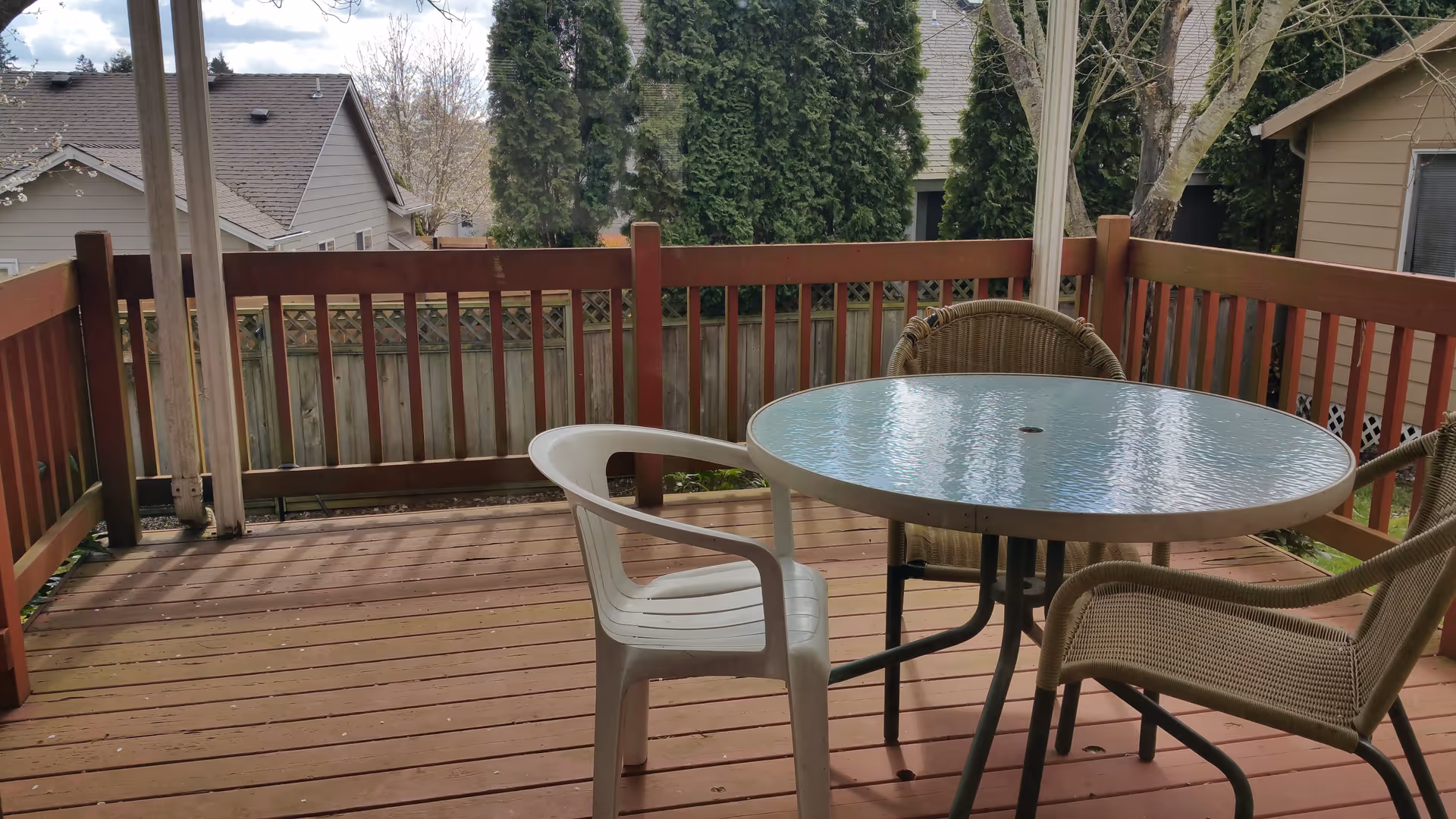 A wooden outdoor deck with a round glass-top table and three chairs, two wicker and one white plastic. The deck is surrounded by a wooden railing and overlooks neighboring houses and tall evergreen trees.