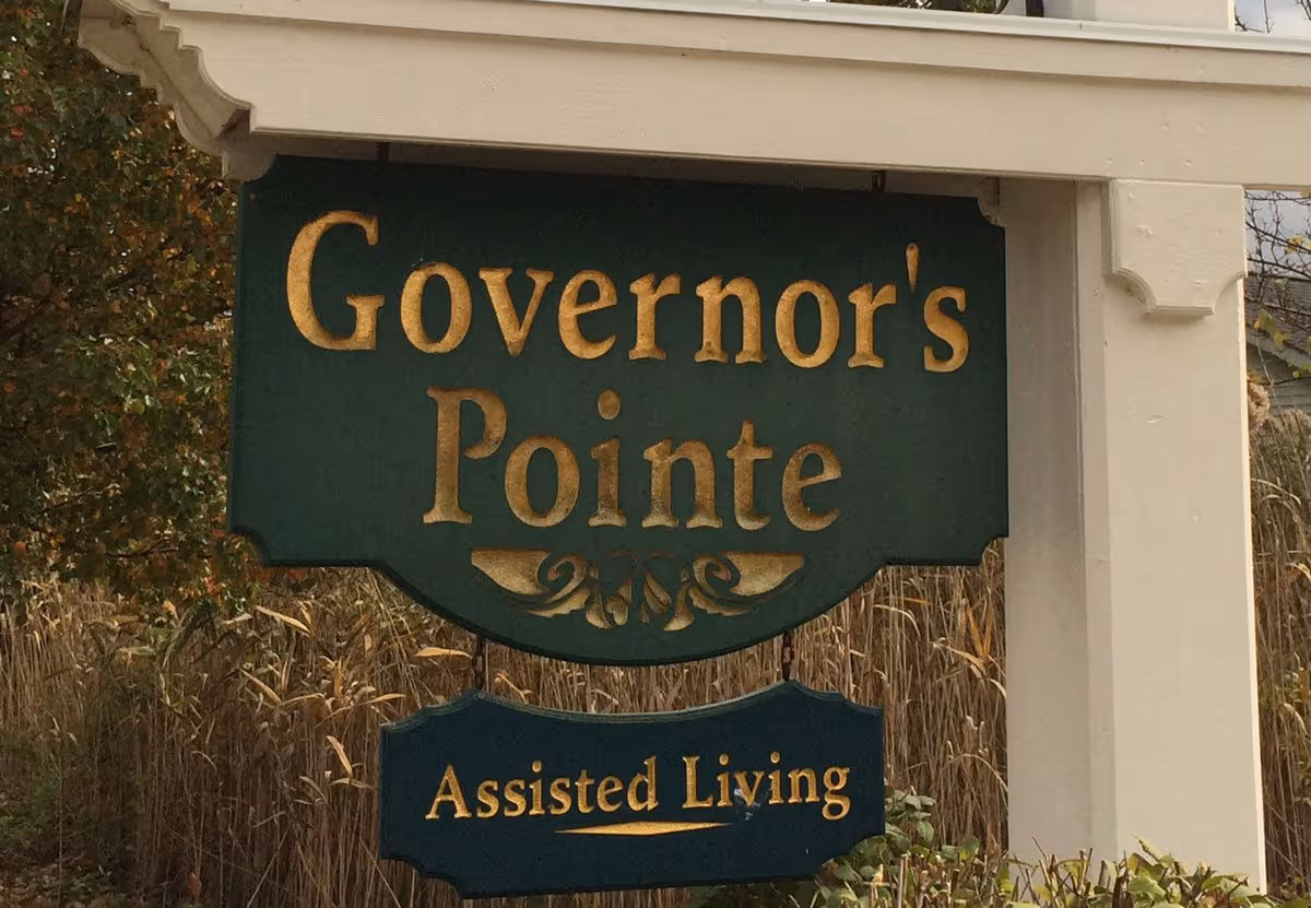 Green wooden sign reading "Governor's Pointe" with a smaller "Assisted Living" sign hanging below on a white post outdoors.