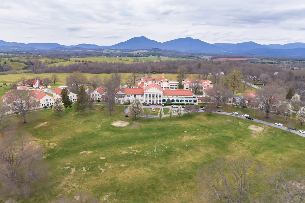 Aerial view of a large senior living facility named Spring Oak at Bedford - The Elks Home, featuring multiple connected buildings with red roofs and white walls, surrounded by expansive green lawns and trees. In the background, there are rolling hills and mountains under a cloudy sky.