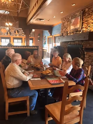 A group of six elderly people sitting around a wooden table in a cozy dining area with stone walls and a fireplace, looking at menus and engaging in conversation.