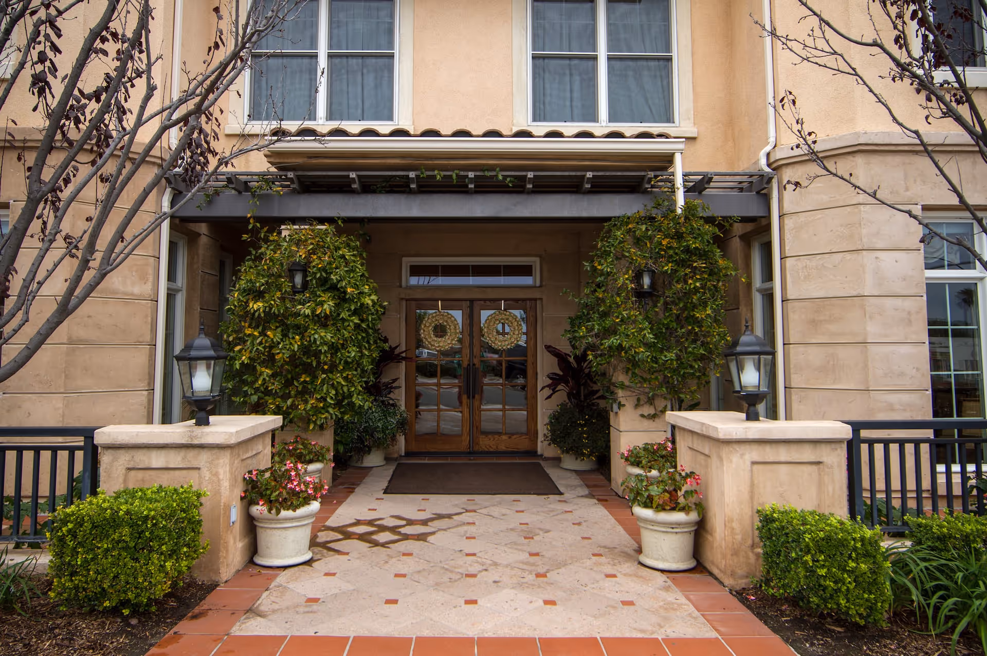 Entrance to a building with double wooden doors decorated with wreaths, flanked by large green plants and potted flowers. The entrance is framed by beige stone walls and has two black lantern-style lights on stone pillars. Leafless trees are visible on either side of the walkway leading to the door.