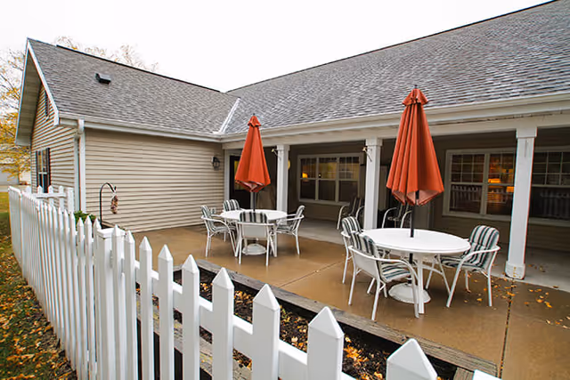 Outdoor patio area with round tables and chairs, each table shaded by a closed orange umbrella. The patio is adjacent to a beige building with a white picket fence in the foreground.