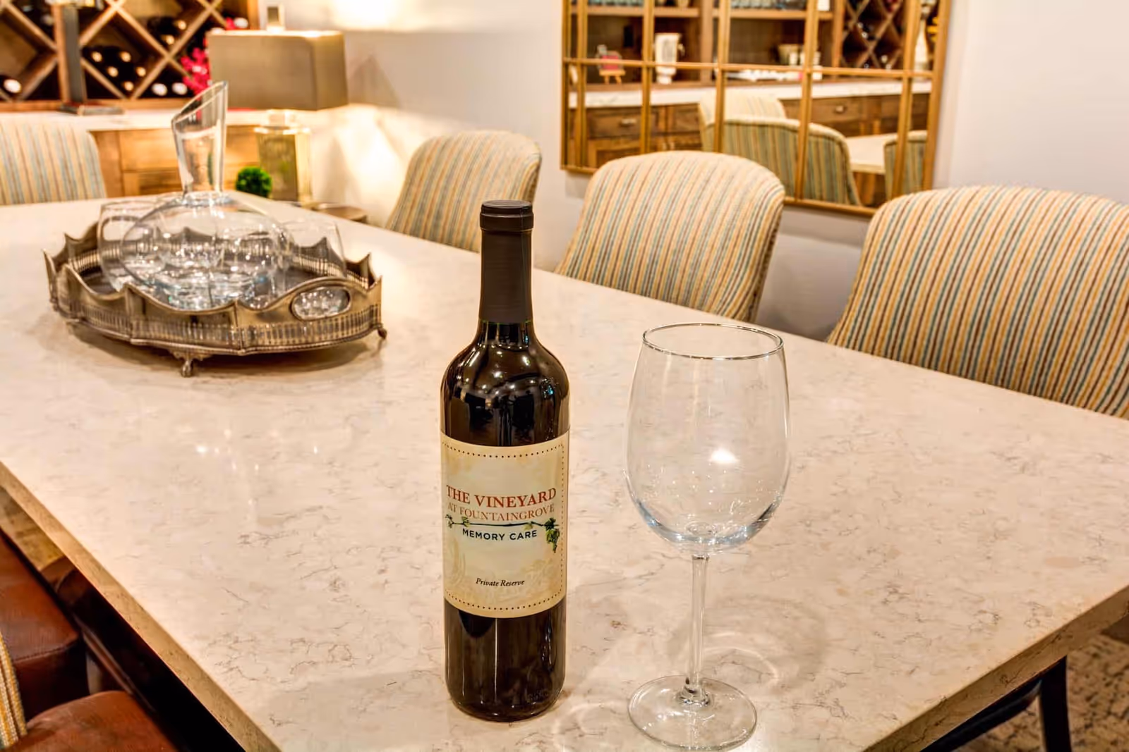 A marble-topped dining island with a bottle of wine labeled "The Vineyard at Fountaingrove Memory Care" and an empty wine glass, with upholstered chairs and wine racks in the background.