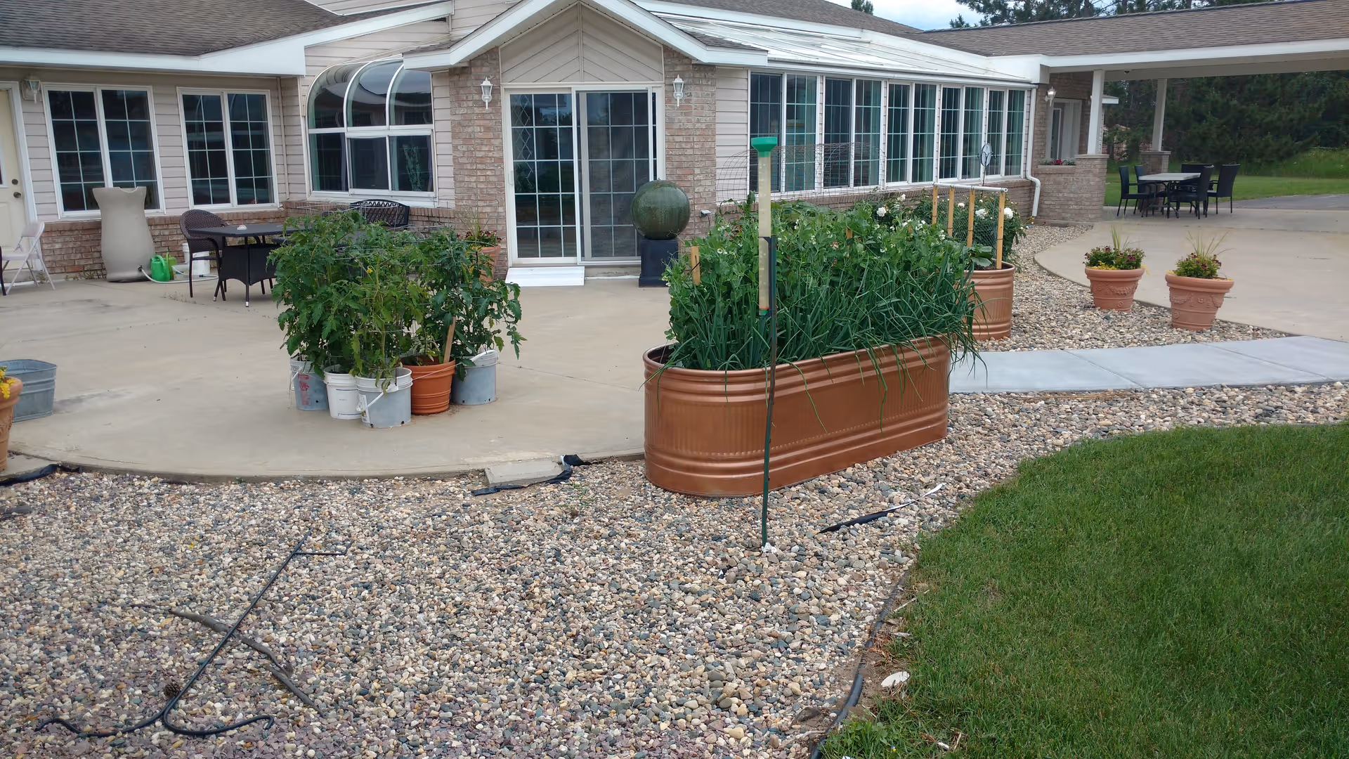 Outdoor patio area of a senior living facility with potted plants and garden beds on a gravel and concrete surface. The building has large windows and a sliding glass door leading to the patio. There are tables and chairs arranged for seating.