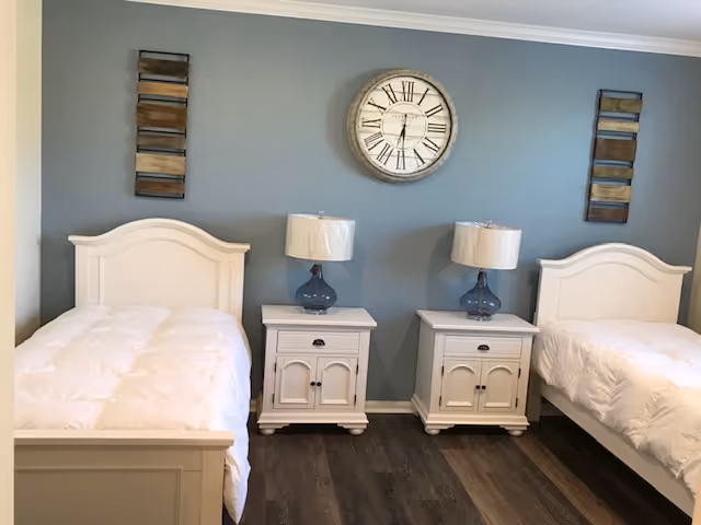 A bedroom with two white twin beds separated by two white nightstands, each with a blue glass lamp. A large round clock with Roman numerals hangs on the blue-gray wall above the nightstands, flanked by two rectangular wooden wall decorations. The floor is dark wood.