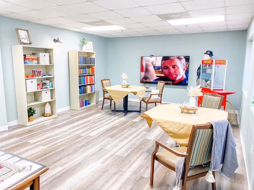 A bright and clean common room with light blue walls and wood flooring. The room features two tables covered with beige tablecloths, each surrounded by chairs. There are two bookshelves filled with books and games against the left wall. A large flat-screen TV is mounted on the far wall, showing a man’s face. To the right of the TV is a vintage-style popcorn machine. The room has a cozy and welcoming atmosphere.