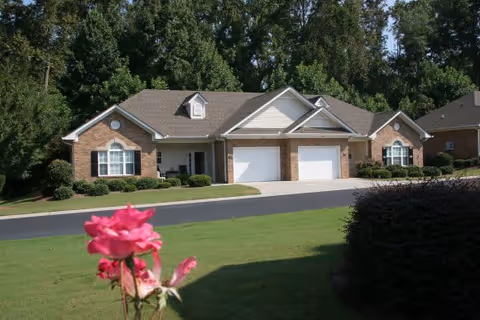 A single-story brick residential building with two white garage doors, surrounded by green grass, bushes, and trees in the background. A pink rose flower is in the foreground on the left side.