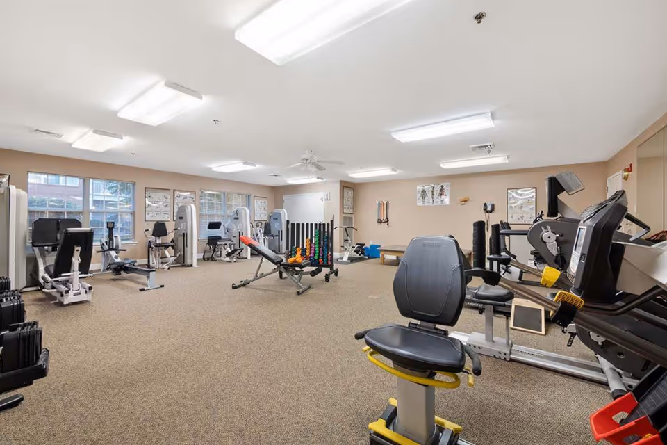 A spacious fitness room with various exercise equipment including stationary bikes, weight machines, dumbbells, and resistance bands. The room has beige walls, carpeted floor, and several windows letting in natural light. Posters related to anatomy and exercise are displayed on the walls.