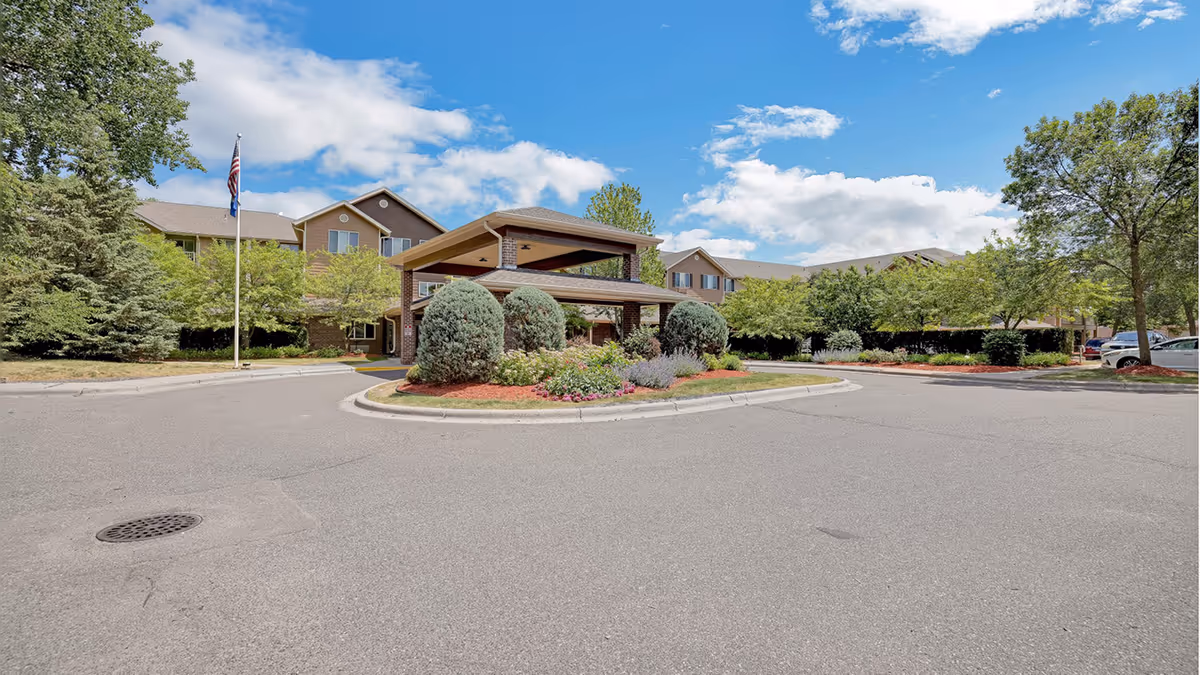 Exterior view of the Lodge At White Bear senior living facility showing the driveway entrance with a covered drop-off area surrounded by landscaped bushes and flowers, trees, and a clear blue sky with some clouds.