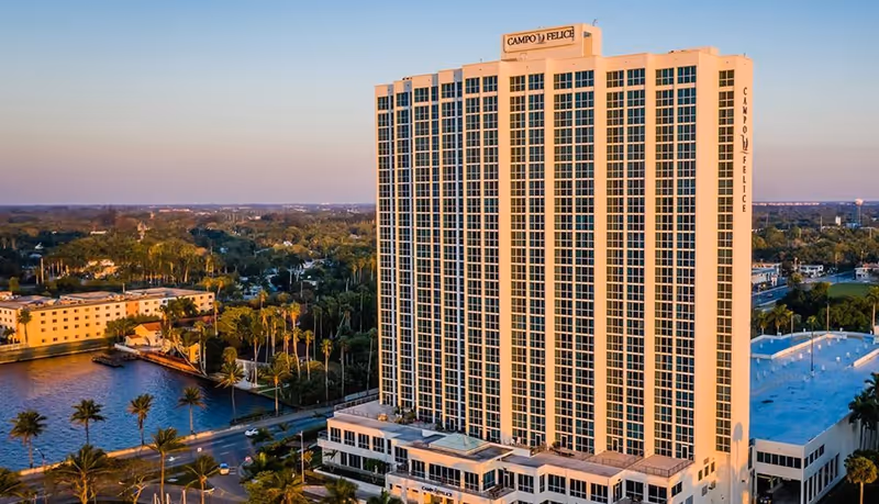 A tall multi-story building named Campo Felice with many windows, surrounded by trees, water, and other buildings under a clear sky at sunset.