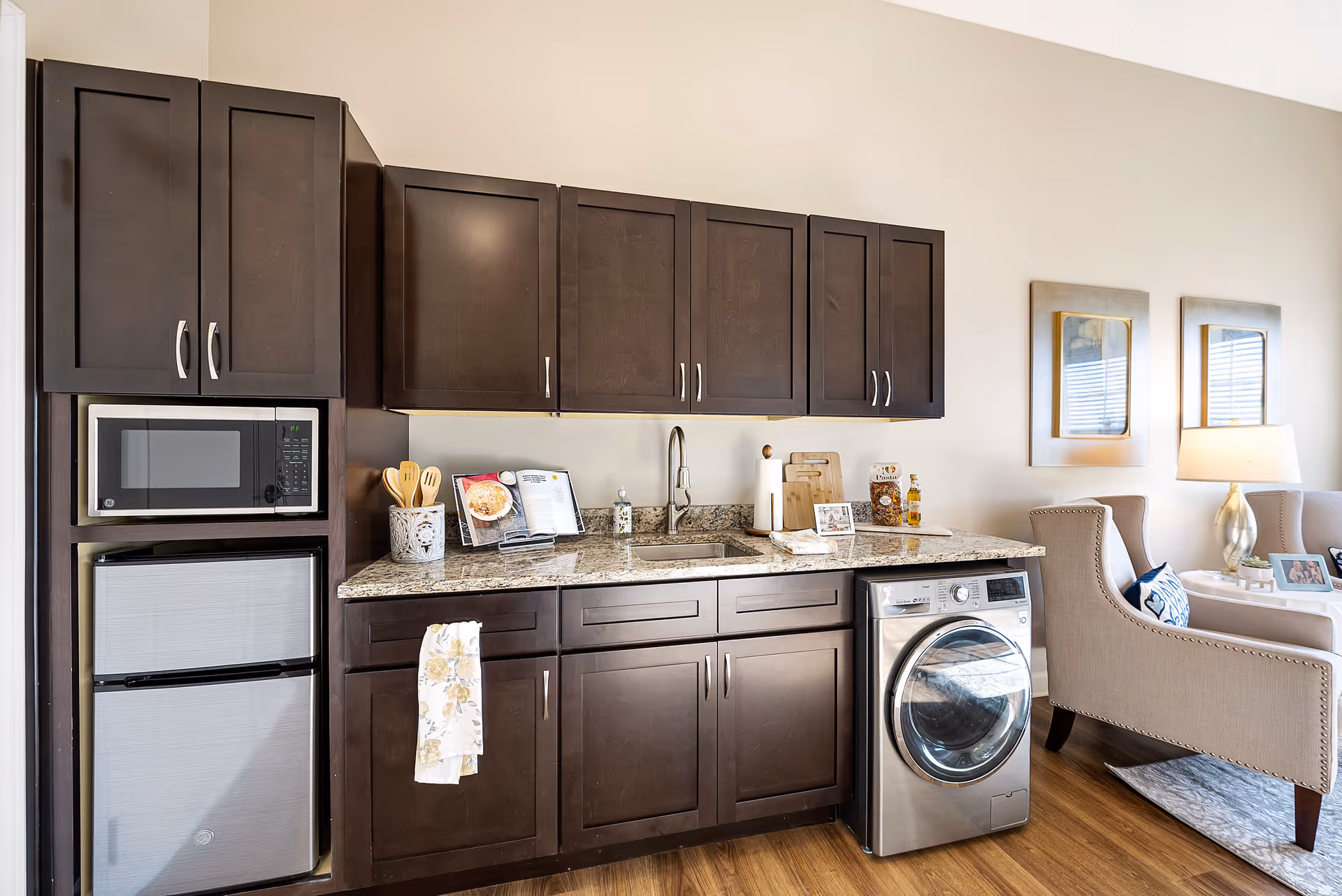 A compact kitchen area with dark wood cabinets, a granite countertop, a microwave above a small refrigerator, a sink, and a front-loading washing machine. To the right, there is a beige armchair and a side table with a lamp and picture frames, with two framed mirrors on the wall behind.