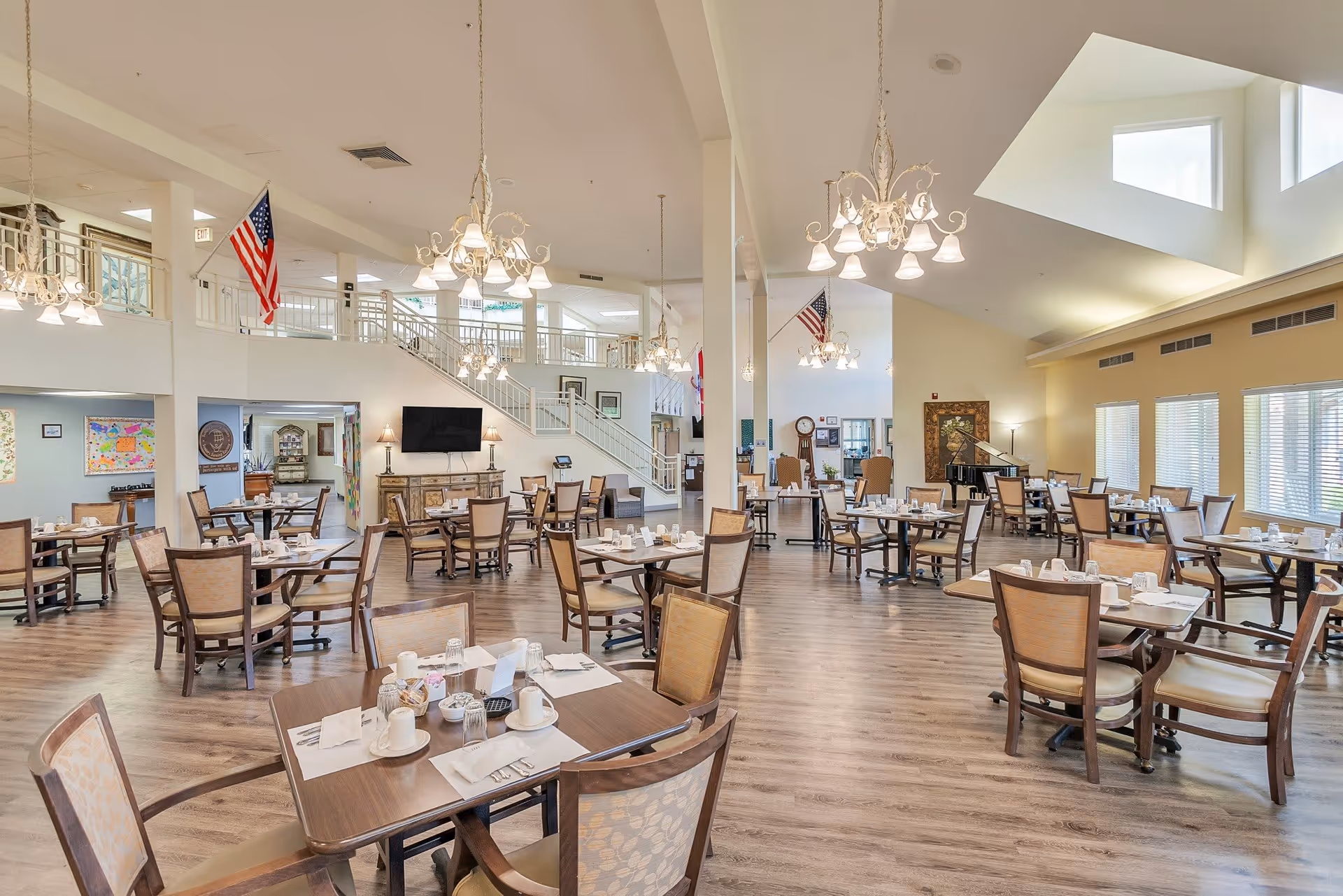 A spacious and well-lit dining room with multiple wooden tables and chairs arranged neatly. Each table is set with white cups, napkins, and utensils. The room features large windows allowing natural light to fill the space, chandeliers hanging from the ceiling, and an American flag displayed near a staircase leading to an upper level.