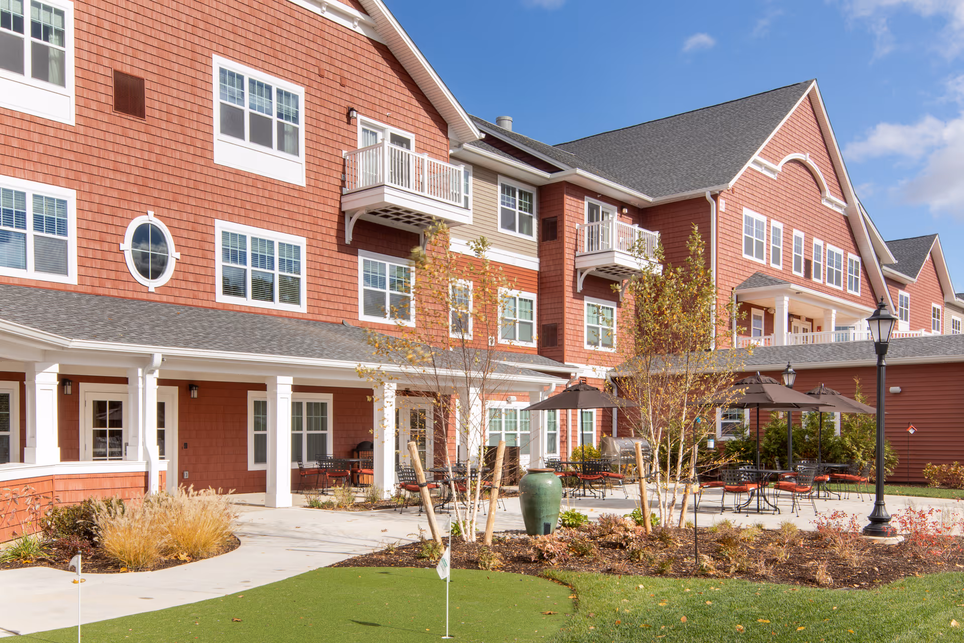Red multi-story senior living building with a landscaped courtyard, outdoor seating and umbrellas, and a small putting green.
