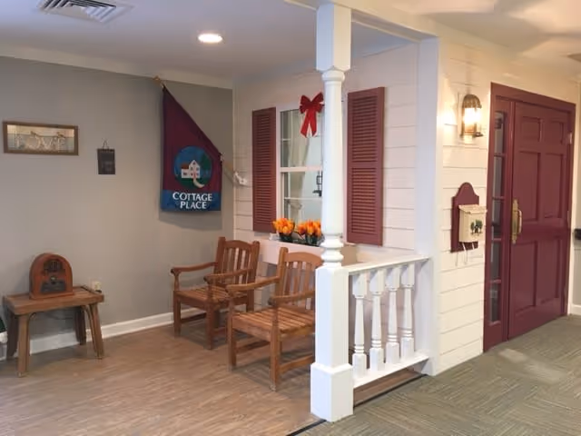 Indoor seating area designed to resemble a porch with two wooden chairs, a small table with a vintage radio, a window with red shutters and a red bow, a flower box with orange flowers, and a maroon door. A flag with the text 'Cottage Place' hangs on the wall.