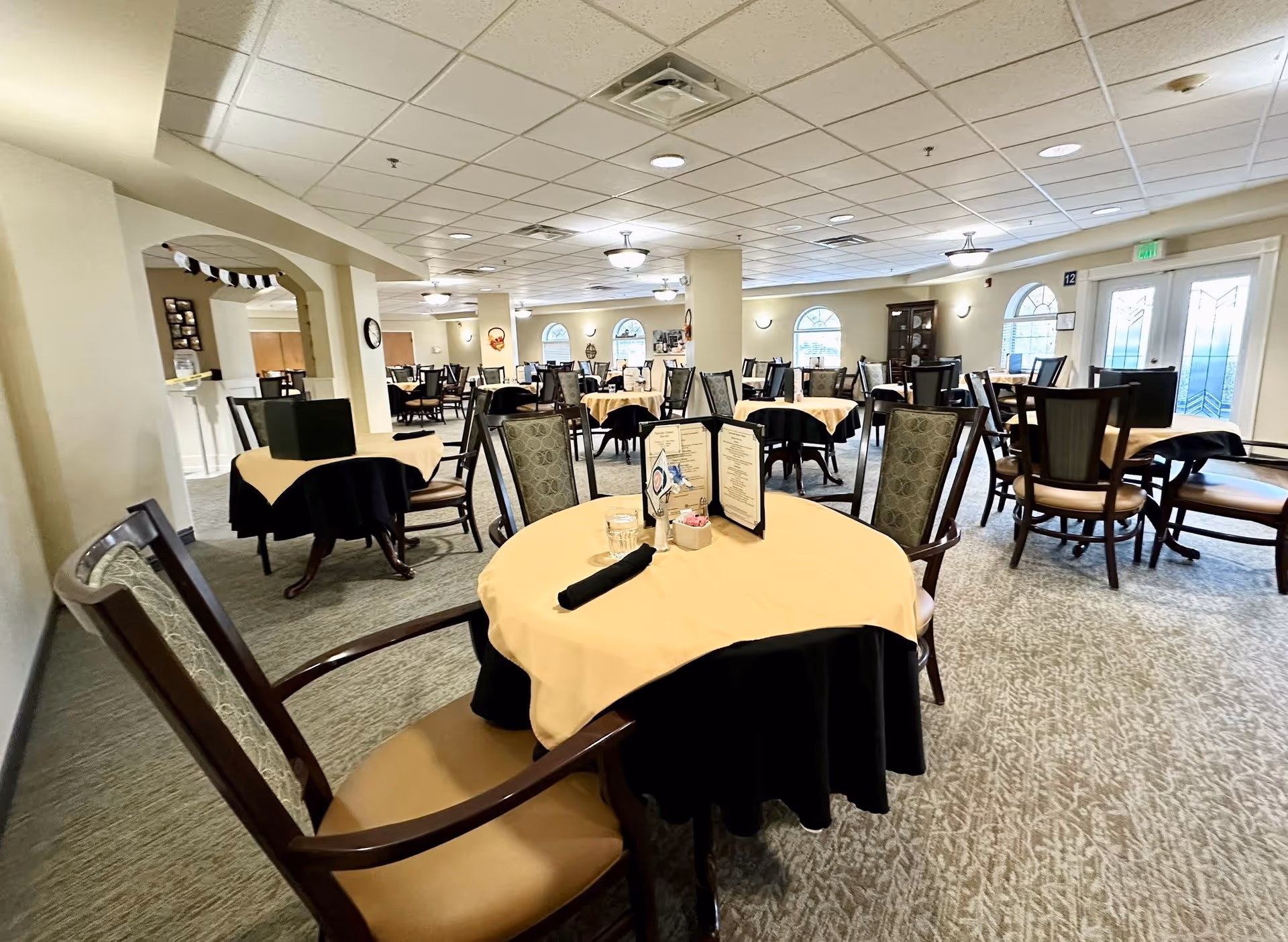 A spacious dining room in a senior living community with multiple round tables covered with yellow and black tablecloths. Each table is surrounded by wooden chairs with cushioned seats and backs. Menus, a glass of water, and a black napkin are placed on the table in the foreground. The room has carpeted floors, beige walls, arched windows, and ceiling lights providing bright illumination.
