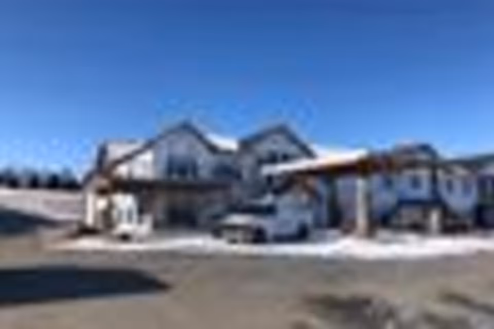 Exterior view of a senior living facility named RiverView Ridge on a clear day with a blue sky. The building has a modern design with multiple gables and a covered entrance where a white vehicle is parked. There is some snow on the ground around the entrance area.