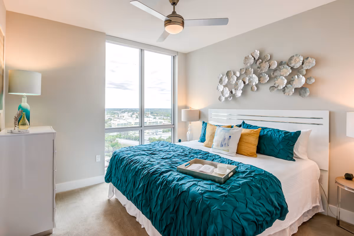 Sunlit modern bedroom with a teal-ruffled bedspread, decorative pillows, wall art above the headboard, bedside lamps, and a large window overlooking the city.