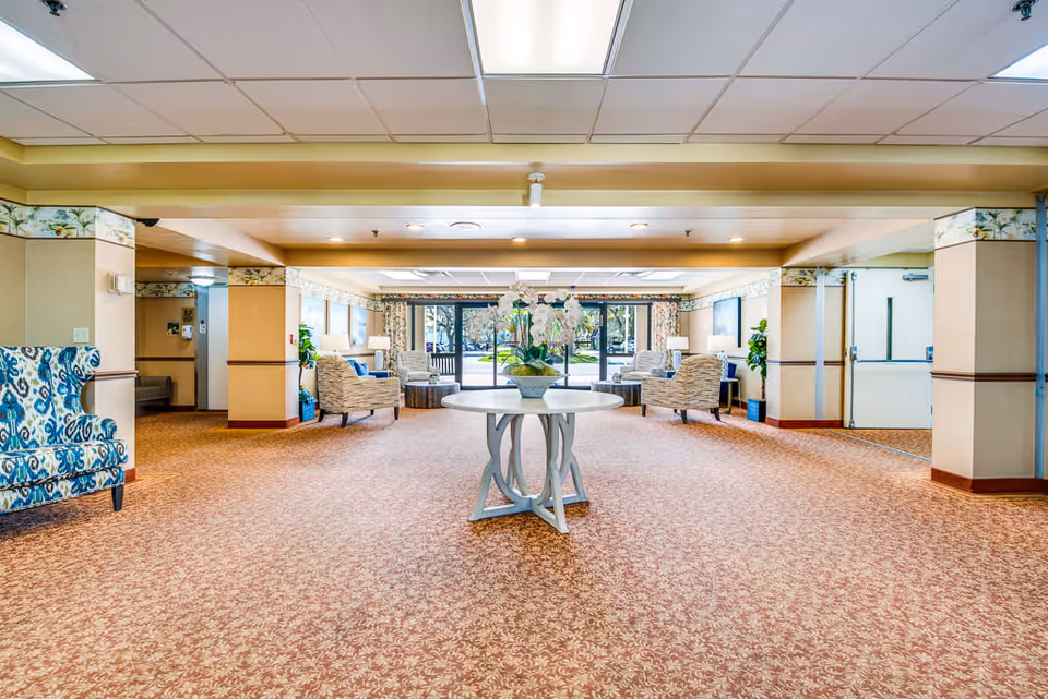 Spacious senior living facility common area with patterned carpet and a round table with a flower arrangement in the center. Comfortable armchairs are arranged near large windows with a view of the outdoor greenery. The room has a drop ceiling with fluorescent lighting and beige walls with decorative trim.