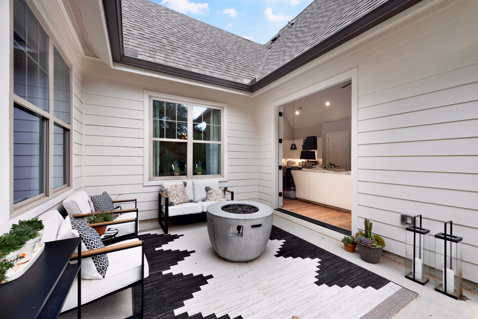 A cozy outdoor patio area with white siding walls, a gray fire pit in the center on a black and white patterned rug, cushioned chairs with decorative pillows, potted plants, and two lanterns. The patio opens into a living room with a white sofa visible through a large open doorway.