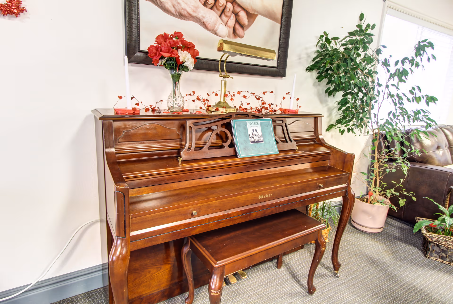 A wooden upright piano with a matching bench in a well-lit room. On top of the piano are two white candles, a vase with red and white flowers, a brass lamp, and a hymns book placed on the music stand. Behind the piano is a framed picture of two hands holding each other. To the right, there is a large potted plant and part of a brown leather couch is visible.