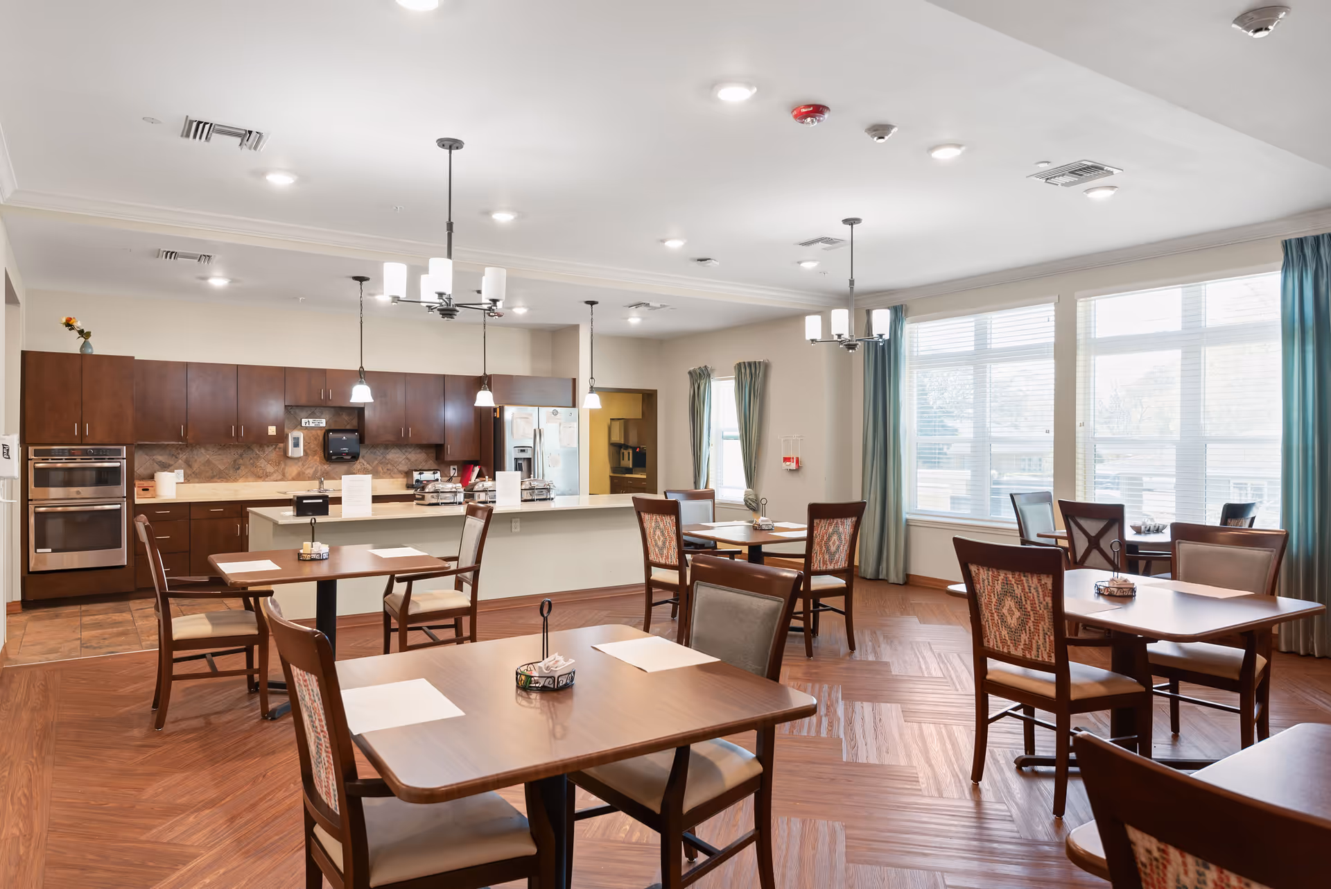 A bright dining room in a senior living facility with several wooden tables and chairs arranged neatly. The room features large windows with teal curtains allowing natural light to fill the space. In the background, there is a kitchen area with dark wood cabinets, a double oven, a refrigerator, and a countertop with pendant lights hanging above.