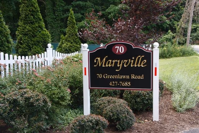 A black and gold sign for Maryville at 70 Greenlawn Road with the phone number 427-7685, surrounded by bushes and greenery with a white picket fence in the background.