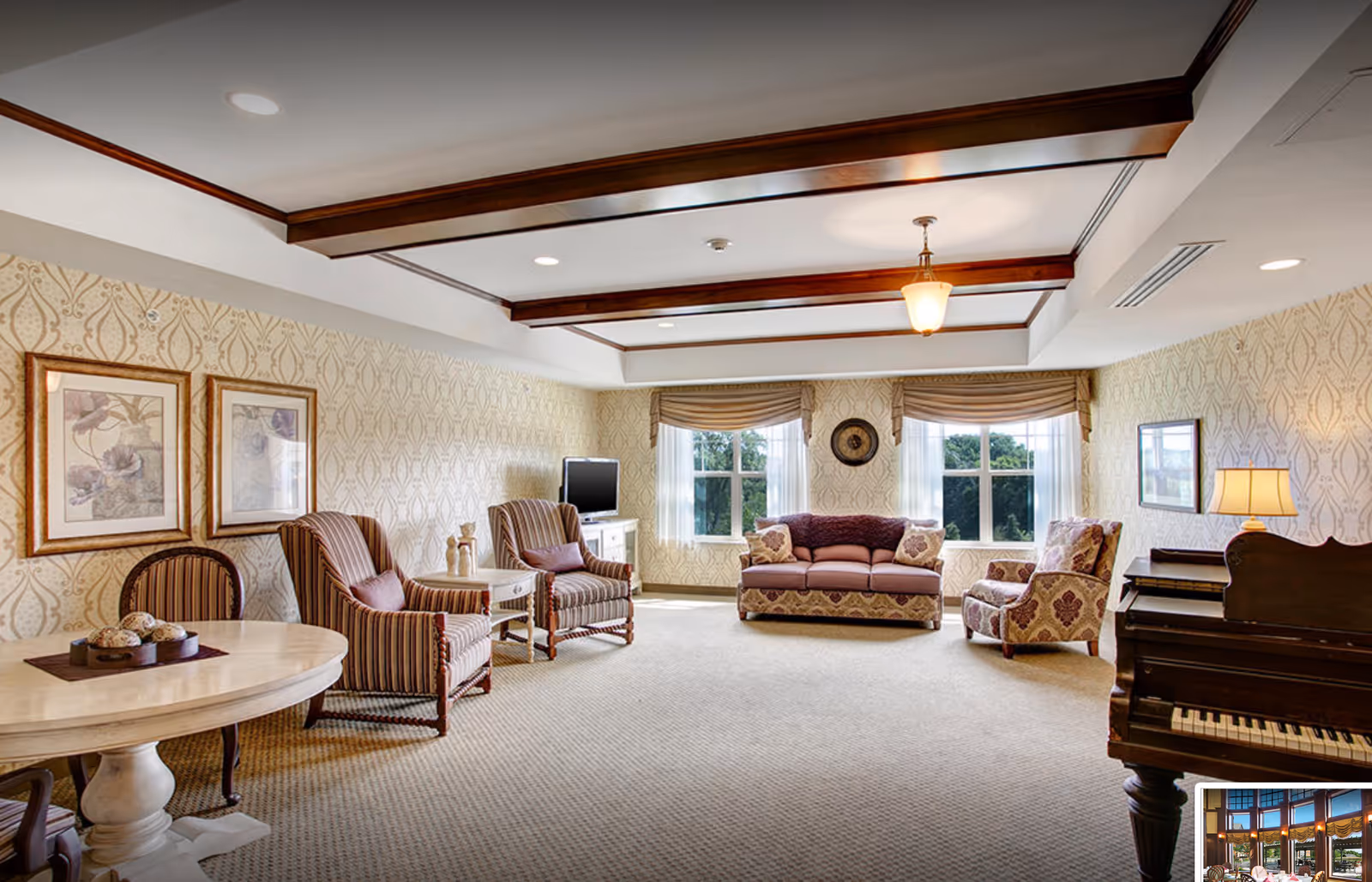 A spacious living room with patterned wallpaper and carpeted floor. The room features a round wooden table with chairs, two striped armchairs with a small side table between them, a flat-screen TV on a white cabinet, a patterned sofa and armchair near two large windows with beige curtains. There is a wooden piano with a lamp on top in the foreground. The ceiling has wooden beams and recessed lighting.