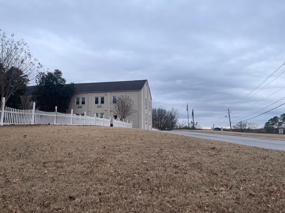 A beige multi-story building sits behind a white picket fence on a grassy slope beside a road under a cloudy sky.