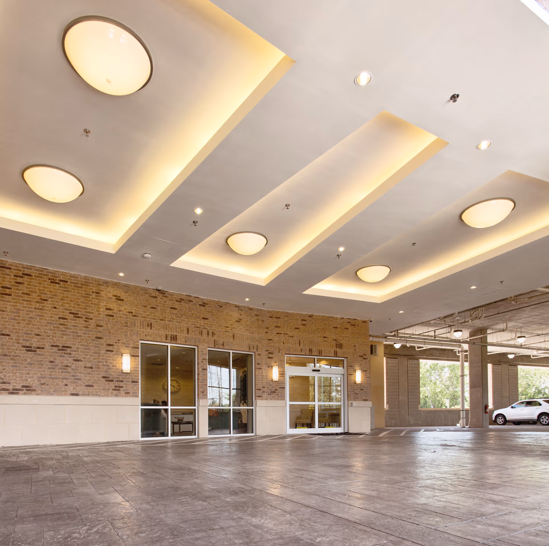 Covered drive-up entrance with recessed ceiling lights, brick facade, glass doors and a parked car visible at the side.