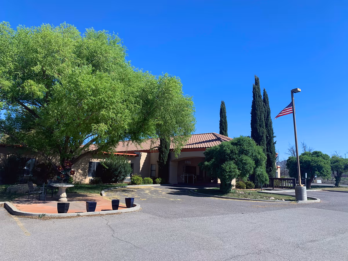 Exterior view of Desert Peaks Assisted Living and Memory Care facility showing a single-story building with a tiled roof, surrounded by green trees and shrubs under a clear blue sky. There is a paved driveway and parking area in front, with an American flag on a tall flagpole to the right.