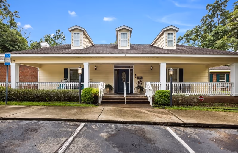 Front exterior view of a single-story building with a covered porch, white railings, and three dormer windows on the roof. There are two lamp posts on either side of the entrance, a wheelchair accessible sign on the left, and a small stop sign on the right. The building is surrounded by greenery and trees under a blue sky with some clouds.