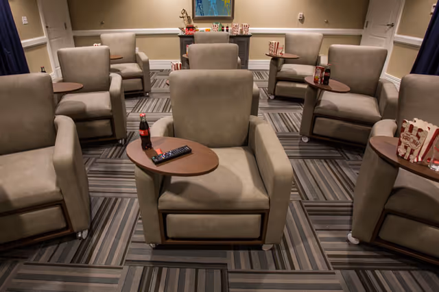 A cozy room with several beige armchairs arranged in rows, each with a small attached wooden table. Some tables have popcorn containers and drinks on them. The room has patterned carpet flooring and neutral-colored walls with a framed picture and a small cabinet at the back.