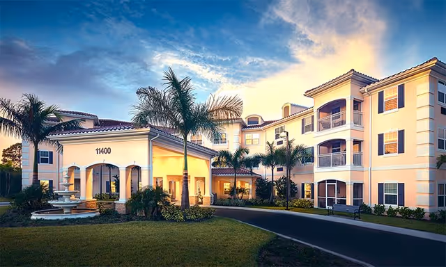 Exterior view of a three-story senior living facility building with a covered entrance, palm trees, a fountain, and a well-maintained lawn under a partly cloudy sky during sunset.